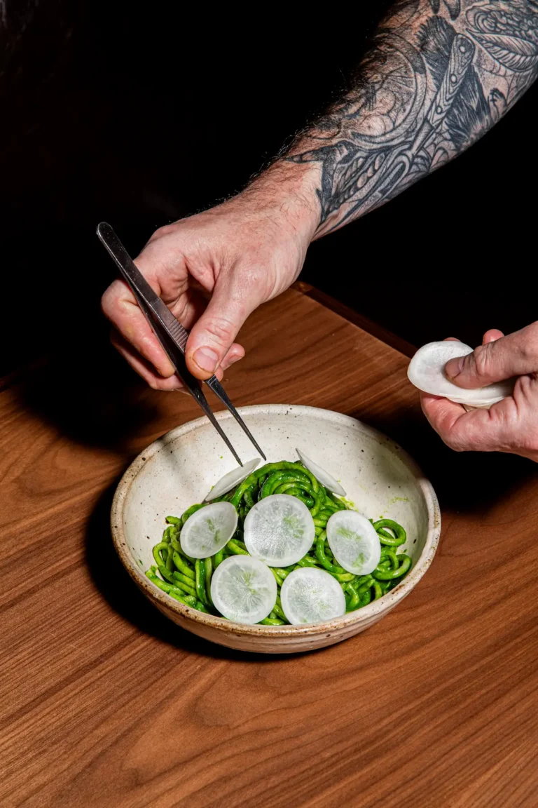 A chef places sliced radish on a dish of green noodels